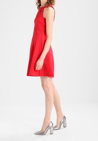 Röd ärmlös klänning med en figursydd bodice och utsvängd kjol, textilerad tyg. Matchas med silverfärgade högklackade skor med blockklack.