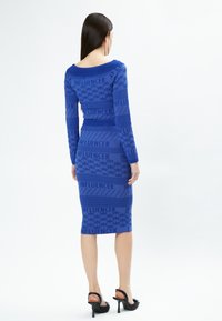 Blauwe bodycon jurk met lange mouwen en een ruime halslijn. Het heeft een gestructureerd patroon met het woord "INFLUENCER" in meerdere stroken.