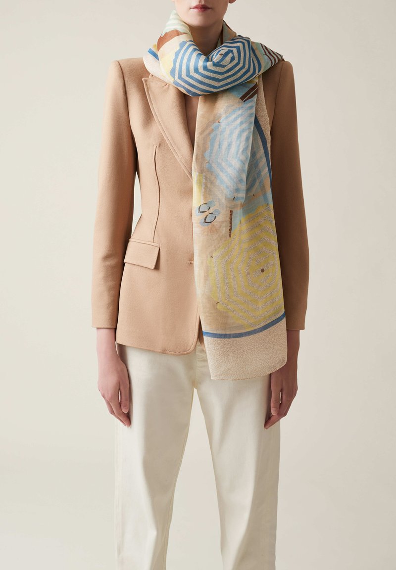 Bedruckter Schal mit geometrischem Muster in Blau, Gelb und Beige, aus weichem Stoff, der um eine Figur in Blazer geschlungen ist.