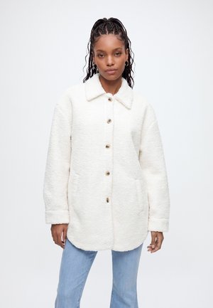 Femme aux cheveux tressés portant des boucles d'oreilles créoles et une veste sherpa blanche boutonnée sur un jean bleu clair, sur un fond uni.