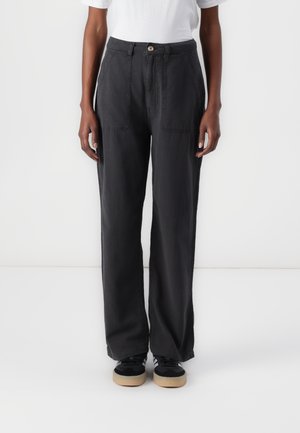 Person iført sorte bukser med vidde ben og forlommer foran, hvid t-shirt og sorte sneakers med beige såler, stående mod en hvid baggrund.