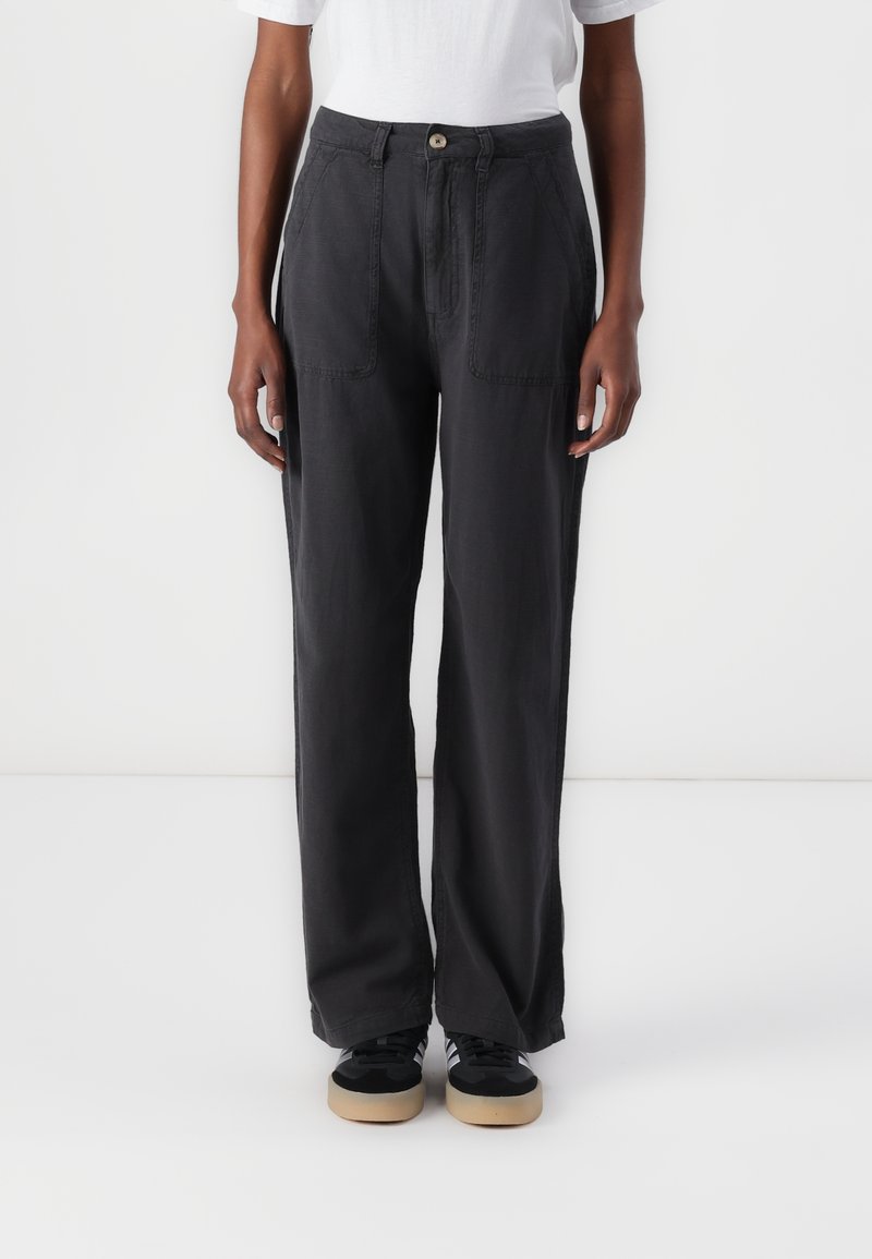 Personne portant un pantalon large noir avec poches plaquées à l'avant, un t-shirt blanc et des baskets noires à semelles beige, debout devant un fond blanc.