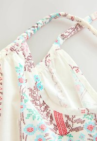 Witte stoffen tanktop met kleurrijke geometrische patronen in roze, blauw en bruin; plooibare schouderbanden en decoratieve stikdetails.