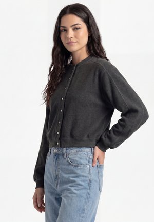 Vrouw met lang donker haar die een donkergebreide vest met knopen en lichtblauwe spijkerbroek draagt, staand met één hand in haar zak.