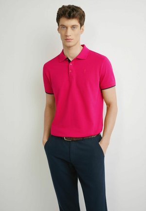 Młody mężczyzna o jasnej karnacji i kręconych brązowych włosach, ubrany w jasnoróżową koszulkę polo i ciemnogranatowe spodnie, stoi oparty o białą ścianę.