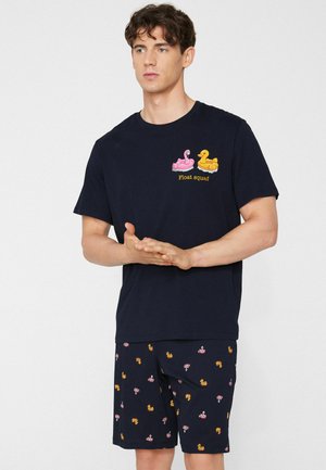 Hombre con camiseta azul marino y pantalones cortos con diseños de flotadores de flamenco rosa y pato amarillo, de pie frente a un fondo blanco liso.