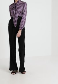 Lila randig blus med knytdetalj och vida ärmar; tillsammans med högmidjade svarta byxor med fickor och sidssprund. Svarta öppna skor.