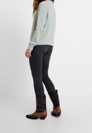 Femme portant un pull en maille gris clair, un jean skinny foncé et des bottes de cowboy noires et marron avec des détails en forme d'étoile, debout devant un fond uni.