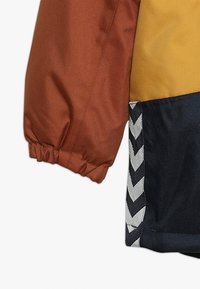 Dettaglio della manica di una giacca color block con polsino elastico arancione ruggine, sezione gialla senape e tessuto blu navy con bordatura a chevron bianca.