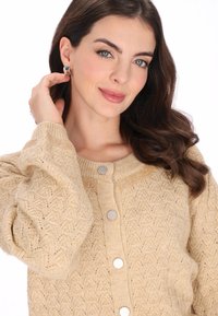 Beżowy sweter z dzianiny o teksturowanym wzorze, okrągłym dekolcie, srebrnymi guzikami i dekoracyjnymi akcentami na ramionach. Miękki materiał.