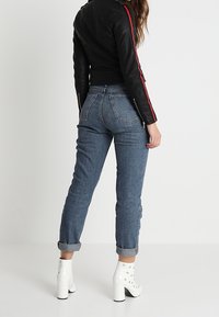 Giacca di pelle nera con strisce rosse, jeans di denim blu arrotolati ai polsini e stivaletti bianchi con tacco alto e ritagli circolari.
