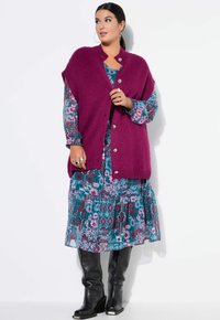 Magenta gestrickte Weste mit silbernen Knöpfen über einem teal- und pinkfarben gemusterten Kleid. Das Outfit hat lange Ärmel und schwarze Stiefel bis zum Knie.
