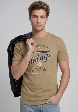 Joven con cabello corto y ondulado lleva una camiseta marrón claro con estampado vintage, una cadena plateada, vaqueros azul claro y sostiene una chaqueta negra sobre su hombro.