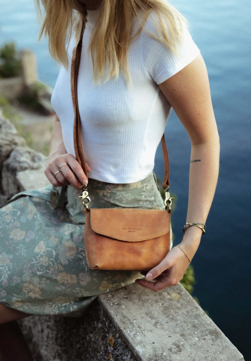 Braune Leder-Crossbody-Tasche mit einem Klappverschluss, verstellbarem Riemen und Metallbeschlägen, die auf dem Schoß einer Person auf einer strukturierten Oberfläche ruht.