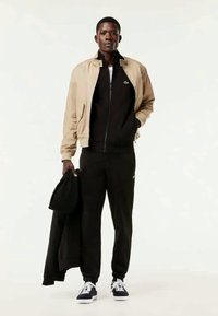 Helle beigefarbene Bomberjacke über einem schwarzen Reißverschluss-Hoodie und schwarzen Jogginghosen. Schwarze Sneakers mit weißen Sohlen. Hält einen schwarzen Sweatshirt.