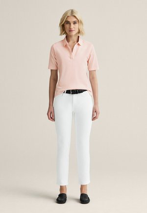 Kvinde stående iført lyserød kortærmet polo, hvide bukser med sort bælte og sorte slip-on sko på en ensfarvet baggrund.