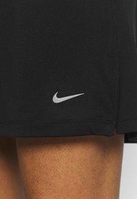 Musta urheiluhame, joka on valmistettu sileästä kankaasta, jossa on kaareva helma ja pieni valkoinen Nike-logo vasemmassa alakulmassa.