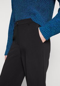 Gros plan d'une personne portant un pull en laine texturé bleu et un pantalon noir, avec une main placée dans la poche du pantalon.