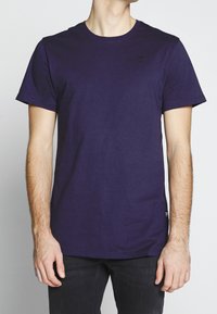 Mies, który ma na sobie gładki ciemnofioletowy t-shirt z krótkim rękawem i czarne spodnie, stoi na tle jasnego, jednolitego tła.