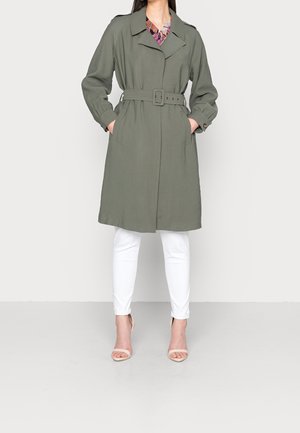 Vrouw die een olijfgroene trenchcoat met ceintuur draagt over een bloemenblouse, witte broek en nude open-teen hakken, staand met handen in de zakken.