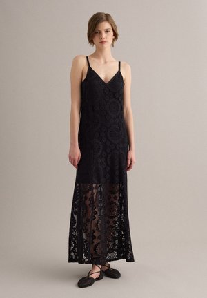 Donna che indossa un abito lungo senza maniche in pizzo nero con spalline sottili e scarpe basse nere intrecciate, in piedi davanti a uno sfondo semplice.