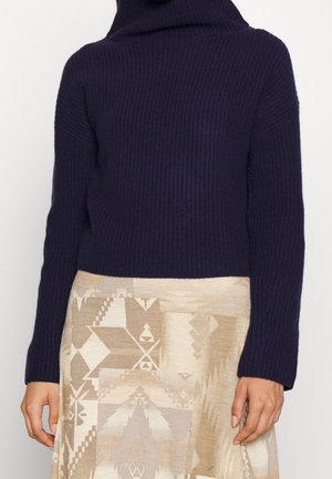 Femme portant un pull à col roulé côtelé bleu marine et une jupe beige à motifs géométriques, debout les bras détendus le long du corps.