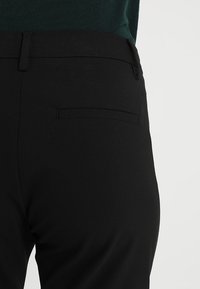 Gros plan sur une personne portant un pantalon noir sur mesure avec une poche arrière passepoilée et un haut vert foncé rentré.