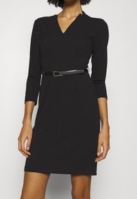Vestido negro con mangas largas, escote en V, corte ajustado y un cinturón negro delgado en la cintura. Tejido suave, diseño hasta la rodilla.