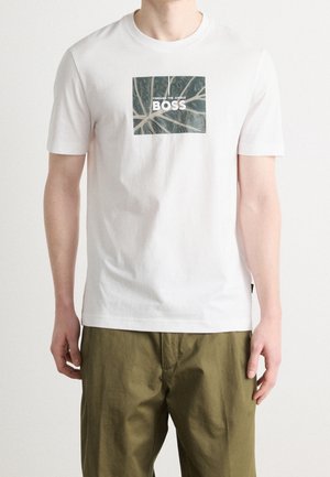 Mężczyzna w białej koszulce z zielonym liściem i napisem „THROUGH THE JUNGLE BOSS”, w połączeniu z oliwkowozielonymi spodniami.