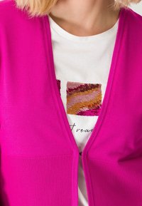 Jasnoróżowy kardigan o prążkowanej fakturze i dekolcie w serek, noszony na białej koszuli z naszytym wzorem z cekinów w odcieniach różu, złota i brązu.