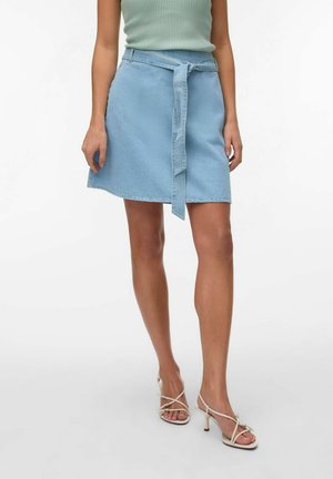 Kobieta ubrana w jasnoniebieską dżinsową spódnicę z przewiązanym paskiem, zielony prążkowany top bez rękawów oraz beżowe sandały na obcasie z paskami.