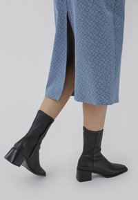 Bottines en cuir noir avec un bout pointu et un talon bloc, associées à une jupe à motif bleu clair avec des fentes sur les côtés.