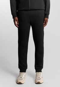Schwarze Jogginghosen aus einem weichen, dehnbaren Material, mit schmal zulaufenden Beinen und gerippten Bündchen. Kombiniert mit hellen Sportschuhen.