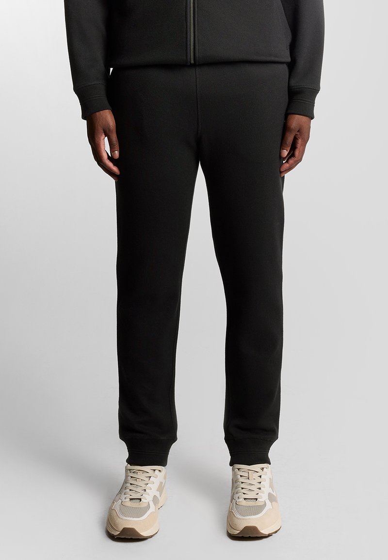 Schwarze Jogginghosen aus einem weichen, dehnbaren Material, mit schmal zulaufenden Beinen und gerippten Bündchen. Kombiniert mit hellen Sportschuhen.