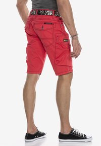 Rote Cargo-Shorts mit mehreren Taschen, sichtbaren Nähten, einem schwarzen Nieten-Gürtel und kombiniert mit schwarzen Sneakers aus Canvas.