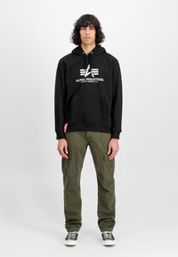 Felpa nera con logo bianco, cappuccio con cordino, tasca kangaroo frontale, abbinata a pantaloni cargo verde oliva e sneaker nere con lacci.
