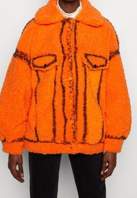 Oranžová nadýchaná bunda s texturovaným, načechraným povrchem. Má černé pruhované detaily a dvě náprsní kapsy. Zaoblený límec a zapínání na knoflíky.