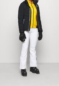 Czarna kurtka puchowa, żółta warstwa pośrednia, białe spodnie narciarskie, czarne rękawiczki i buty narciarskie z różowymi akcentami. Smukły design z kontrastującymi kolorami.