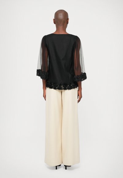 Black sheer top with a floral lace hem, paired with wide beige trousers. The top features semi-transparent sleeves and a round neckline.