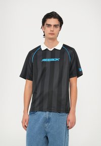 Melnām svītrām Reebok polo krekls ar zilu logotipu, baltu apkakli, īsām piedurknēm un brīvu piegriezumu. Nēsāts ar gaiši ziliem džinsu biksēm.