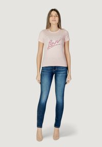 Camiseta de color rosa claro con la inscripción "GUESS LOVE" en texto brillante, combinada con jeans ajustados de color azul oscuro y tacones nude. Tela suave, escote redondo.