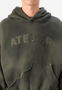 Olivengrønn hettegenser med et slitt "ATELIER"-logo, frontlomme og synlige syninger. Myk stoff med avslappet passform.