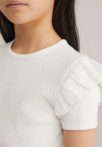 Maglia bianca a maniche corte a coste con morbide balze sovrapposte su una spalla. Il tessuto ha una texture sottile e un design aderente.