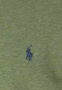 Zielona tkanina o teksturze z małym, granatowym haftowanym logiem przedstawiającym gracza polo na koniu, który macha kijem.