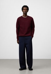 Burgunderfarbener Strickpullover mit Rundhalsausschnitt, kombiniert mit weiten navyblauen Hosen. Das Model steht vor einem schlichten weißen Hintergrund. Schwarze Schuhe.