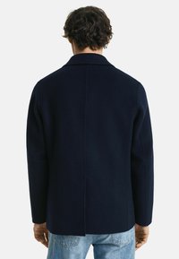 Marineblauwe wollen coat met een rechte snit, kraag en een naad in het midden van de rug. Standaard lange mouwen en minimale afwerking. Gecombineerd met lichtblauwe jeans.