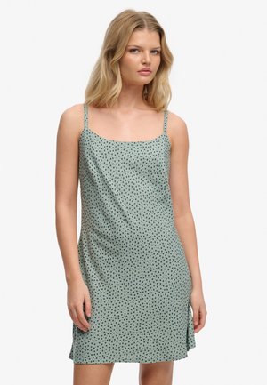 Lyshåret kvinde iført en lysegrøn ærmeløs kjole med tynde stropper og et lille, mørkt blomsterprint, stående foran en ensfarvet baggrund.