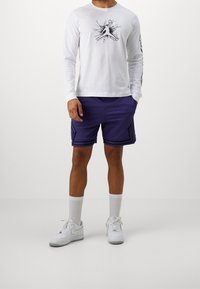 Pitkähihaiset valkoiset paidat mustalla graafisella kuviolla, yhdistettynä violetteihin urheilushortseihin, joissa on mustia yksityiskohtia, sekä valkoisiin tennareihin ja sukkiin.