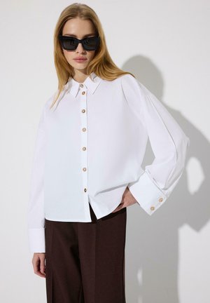 Žena v nadrozmernej bielej košeli na gombíky, tmavých širokých nohaviciach a čiernych nadrozmerných slnečných okuliaroch pózuje na bielom pozadí.