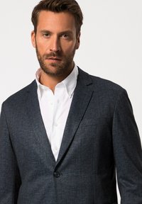 Blazer blu scuro in tessuto strutturato, chiusura con un bottone singolo, revers a lancia, abbinato a una camicia bianca con colletto sotto.